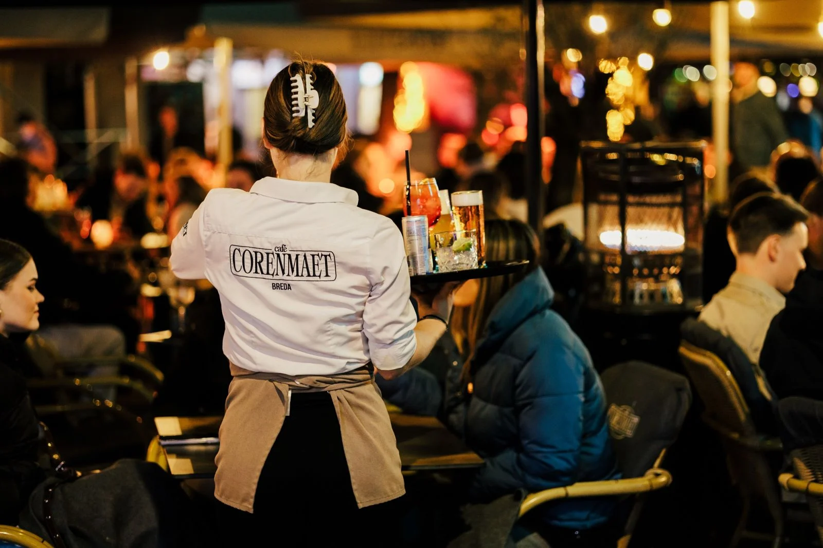 De rug van een medewerker van het Café Corenmaet Breda met het logo is zichtbaar. De medewerkster loopt op het terras met een dienblad vol glazen.