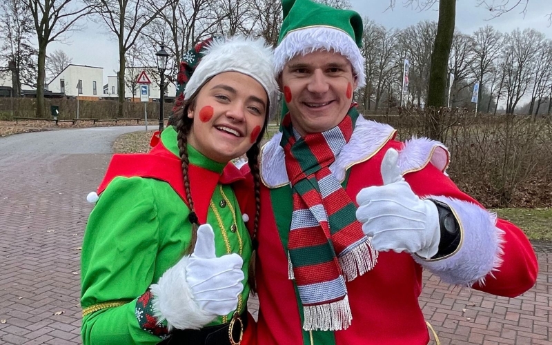 Jordy en zijn collega poseren enthousiast tijdens de kerstparade bij vakantiepark De Leistert. Ze hebben een kerstoutfit aan inclusief kerstmuts en rode geschminkte wangen.