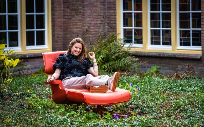 Fem zit buiten in een rode loungestoel in het midden van een groene omgeving. Dit is de binnenplaats van De Rooi Pannen Tilburg Horeca.