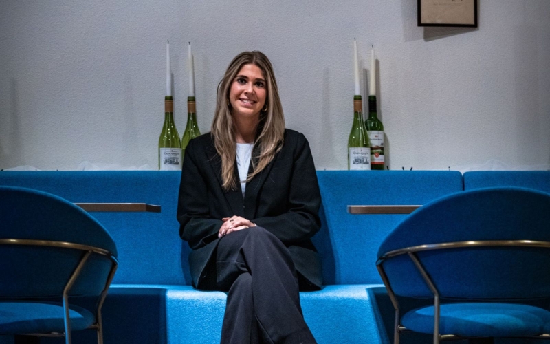 Day zit op een blauw bankje in één van de restaurants bij De Rooi Pannen Tilburg Horeca. Achter haar staan lege flessen wijn met daarin kaarsen. Ze lacht naar de camera.