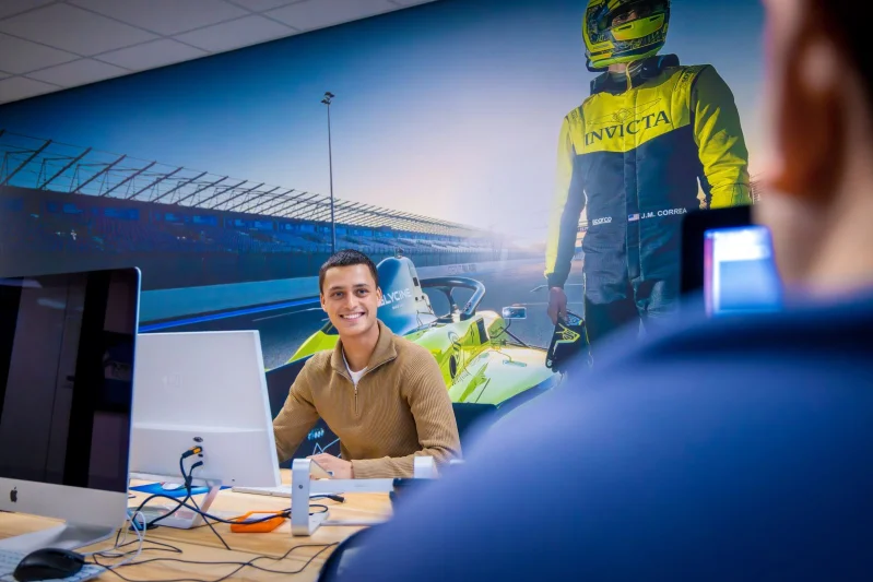 Een student van de mbo-opleiding International Business zit tijdens zijn stage op kantoor, achter een computer en lacht naar een collega tegenover hem.
