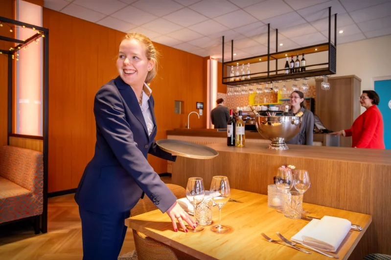 Student Gastheer/gastvrouw in gesprek met twee gasten aan tafel in het restaurant van De Rooi Pannen.