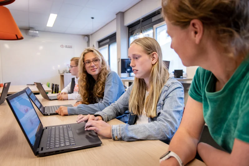 Documenten van De Rooi Pannen Tilburg Vmbo Drie leerlingen van het vmbo van De Rooi Pannen zitten op een rijtje achter hun eigen laptop. Een docent staat naast ze en helpt hen.