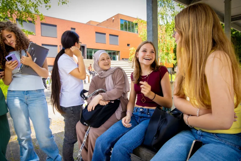 Vakanties voor vmbo bij De Rooi Pannen Een groep leerlingen van het vmbo zitten buiten op een rij en zijn met elkaar aan het kletsen en lachen.